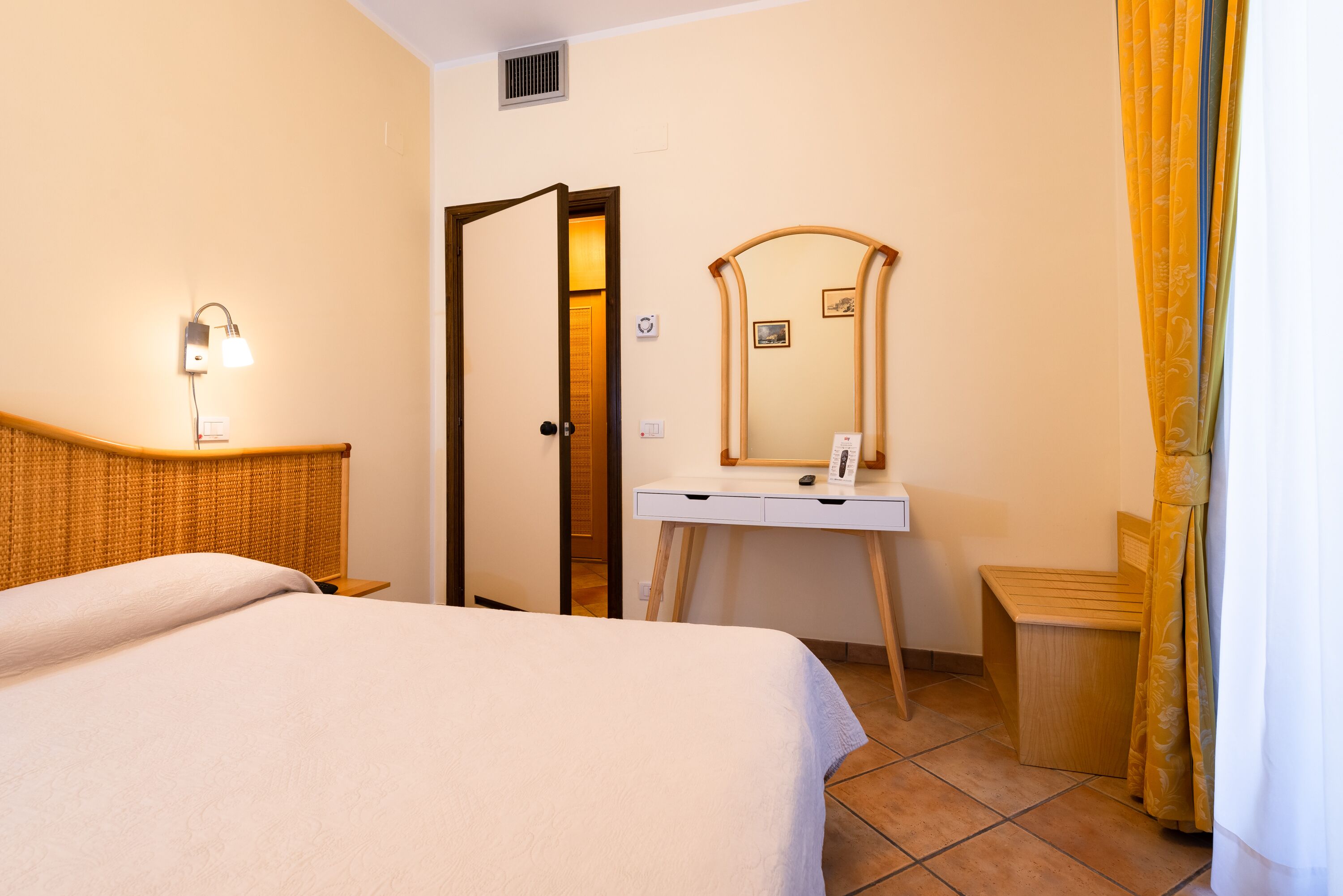 A hotel room with a bed, a bedside table, a desk with a mirror, and a shower area.