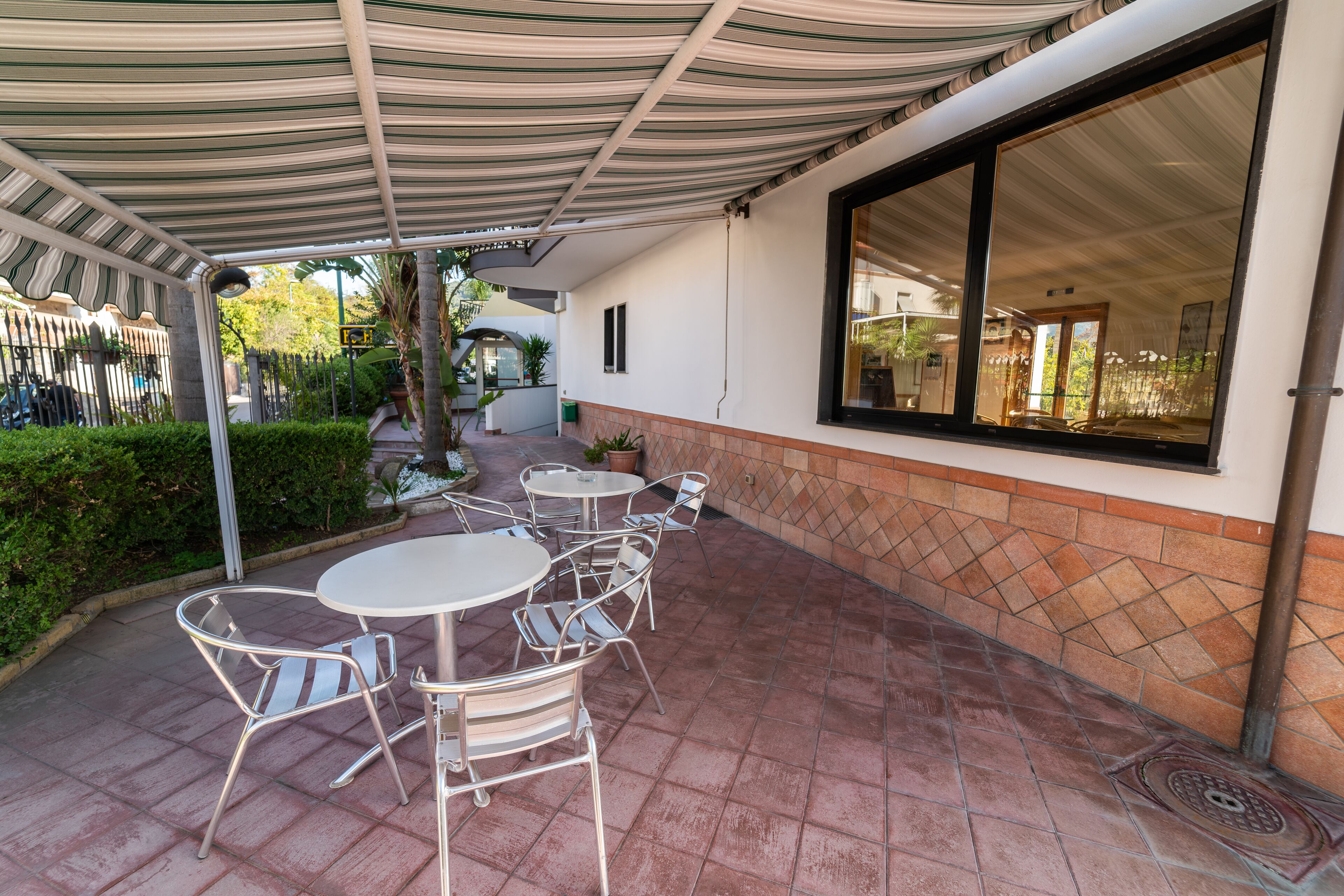Una terrazza esterna con un tenda a righe, tavoli bianchi rotondi e sedie in metallo.