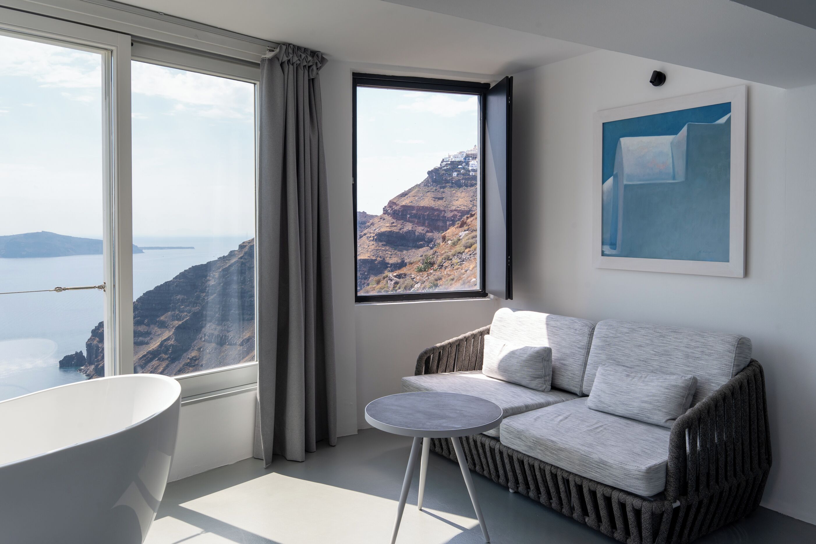 A room with a view of a cliff and sea, a white bathtub, a grey sofa, and a small round table.