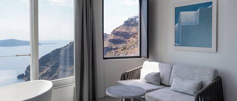 A room with a view of a cliff and sea, a white bathtub, a grey sofa, and a small round table.