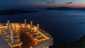 A deck with sun loungers and umbrellas, overlooking a calm sea and distant lights. - Sun Rocks Boutique Hotel | Caldera View Suites & Sunset Dining Experience (Santorini)