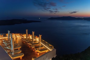 Eine Terrasse mit Liegestühlen und Sonnenschirmen mit Blick auf ein ruhiges Meer und entfernte Lichter.