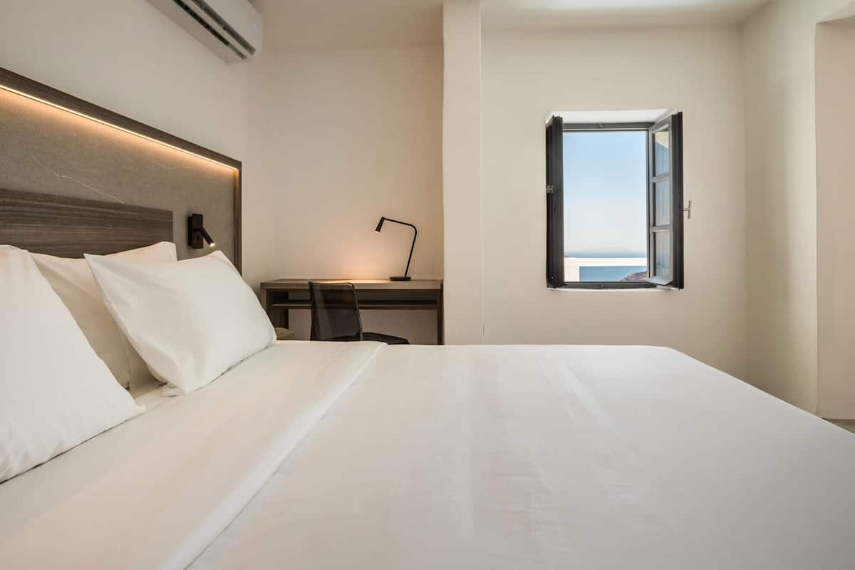 A hotel room with a large bed, a desk with a chair, a window with a view, and a wall-mounted air conditioner.