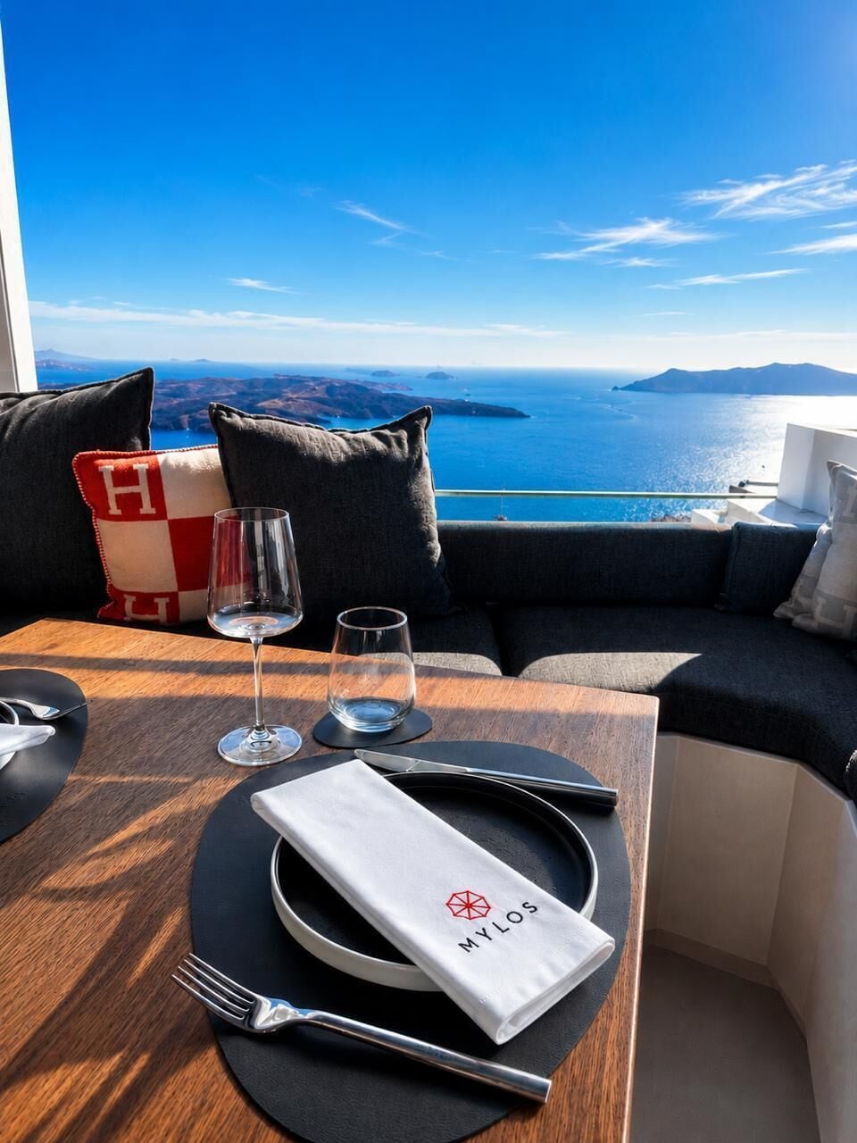 A table set with a napkin, fork, and wine glass, overlooking a scenic view of the sea and islands.