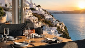 A table set for dining with a view of a coastal town and the sea. - Sun Rocks Boutique Hotel | Caldera View Suites & Sunset Dining Experience (Santorini)