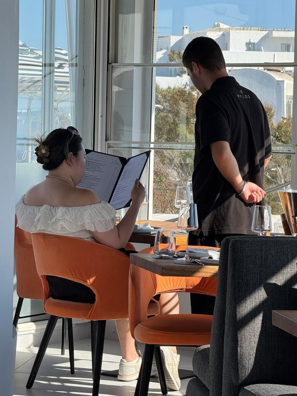 En person sidder ved et bord med en menu, en anden person står ved siden af, og der er udsigt til bygninger gennem store vinduer.