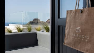 A modern hotel room with a large glass door leading to a balcony with a view of the sea and a rocky outcrop. A brown tote bag hangs on the door frame.