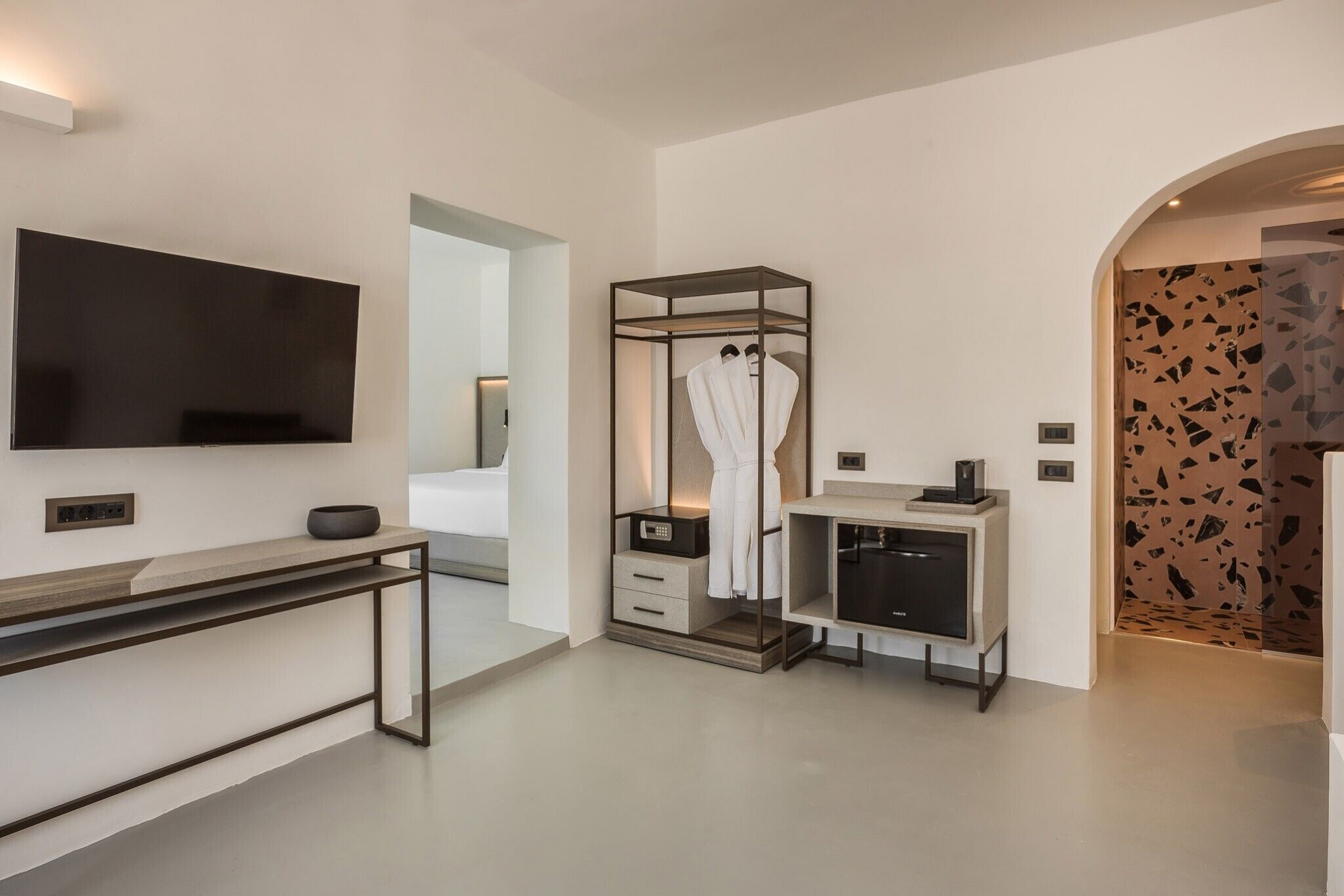 A modern hotel room with a flat-screen TV, a console table with a bowl, a robe closet, a minibar, and a unique wall with a mosaic pattern.