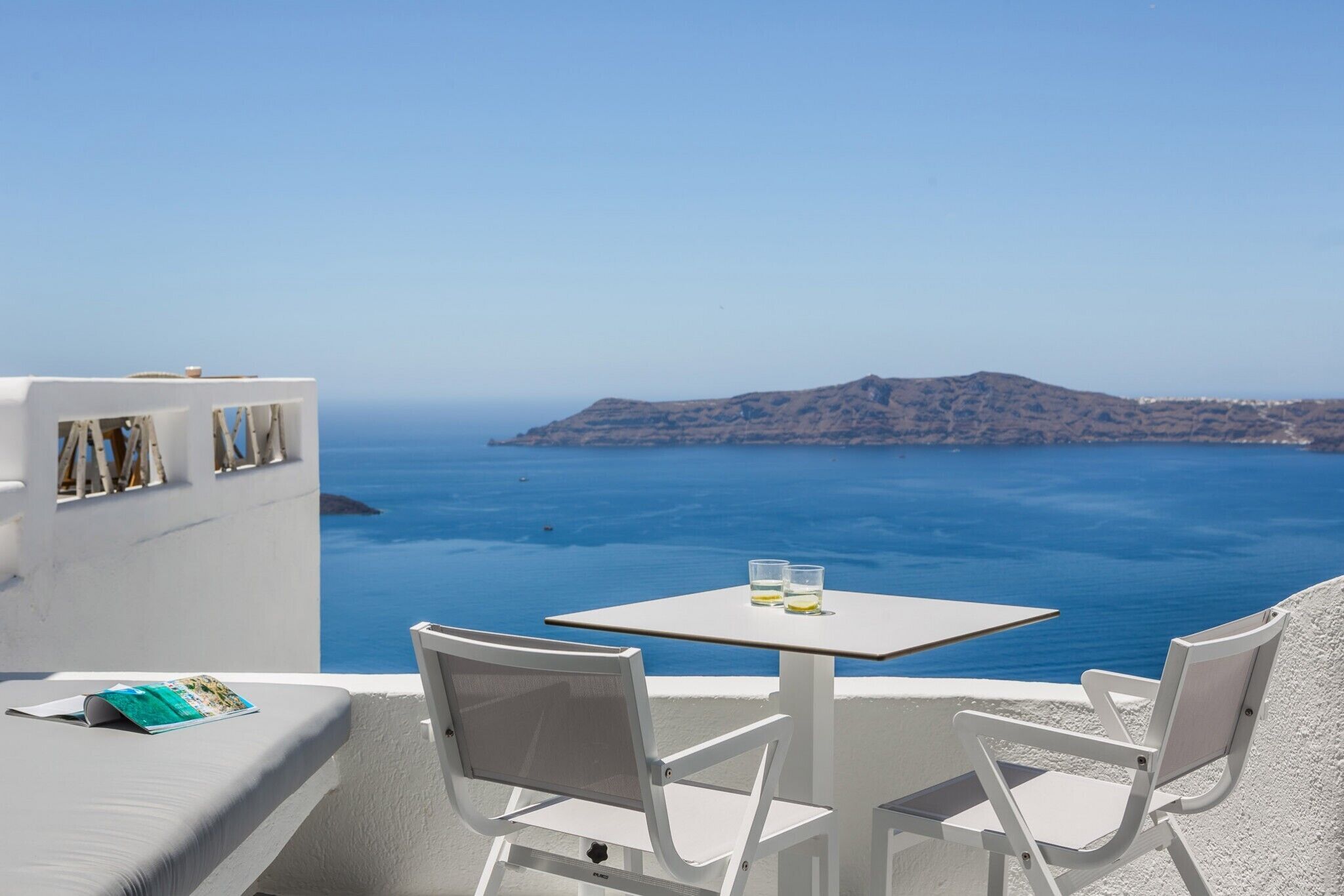 A white outdoor seating area with a table and chairs, overlooking a clear blue sea and a distant island.