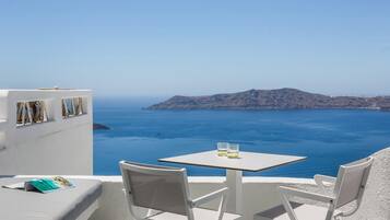 A white outdoor seating area with a table and chairs, overlooking a clear blue sea and a distant island.