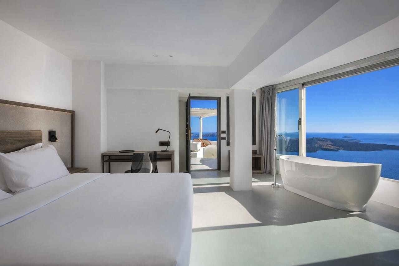 A modern hotel room with a large bed, a desk, a bathroom with a bathtub, and a balcony with a sea view.