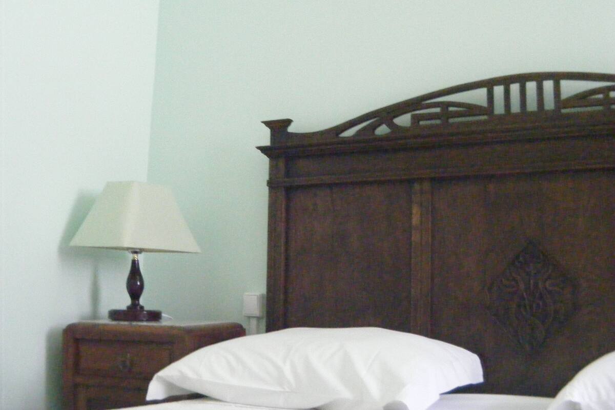 A wooden bed with a headboard, a bedside table with a lamp, and a wall with a light switch.