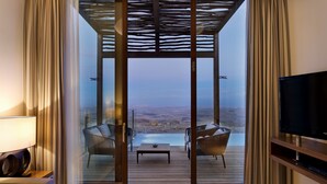 A modern hotel room with a large sliding glass door leading to a balcony with a pool and a view of the desert. There is a flat-screen TV on a wooden stand, a sofa, and a small table with a bowl. - Beresheet by Isrotel exclusive (Ramat Negev)