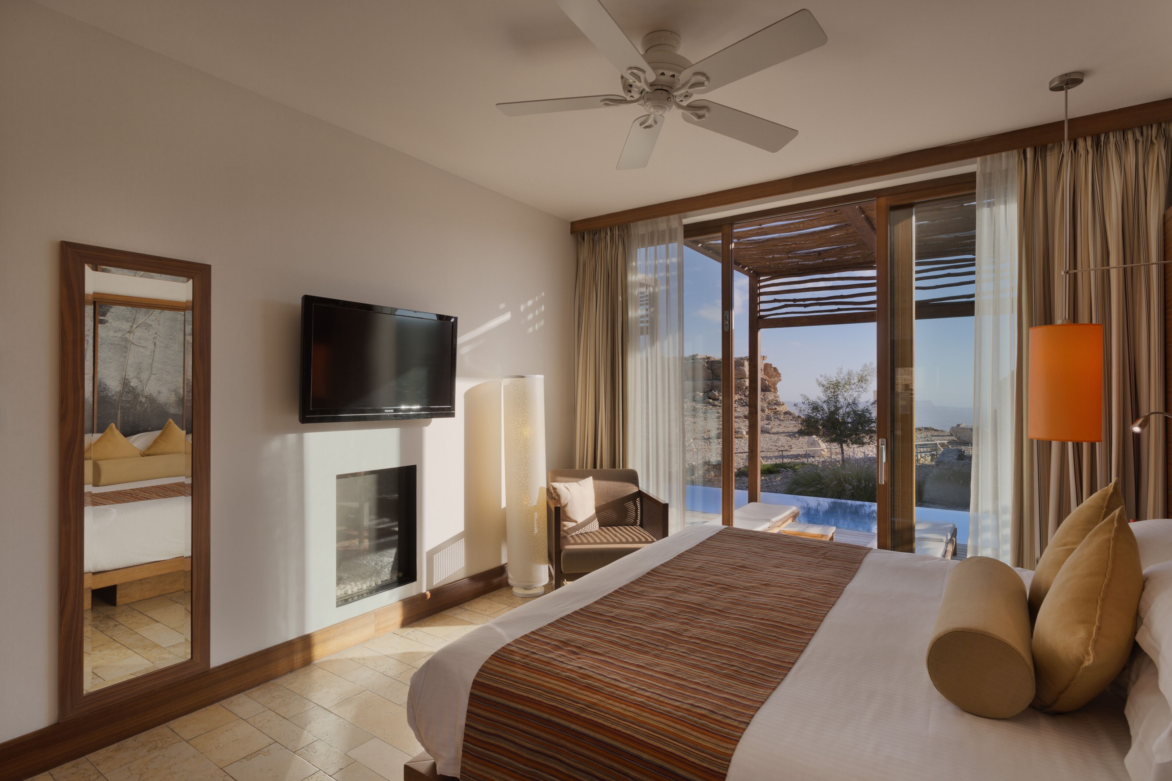 A hotel room with a large bed, a flat-screen TV, a fireplace, a balcony with a view of the landscape, and a ceiling fan.