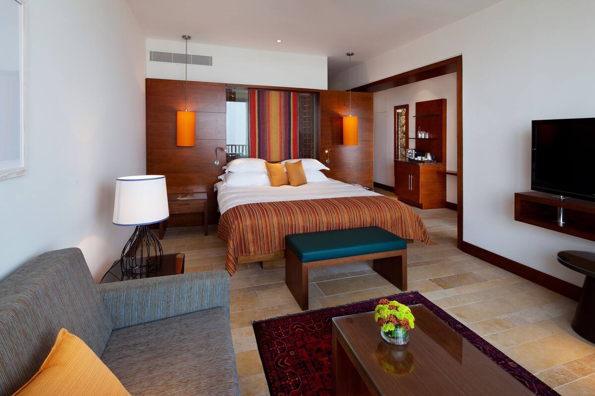 Quarto de hotel moderno com uma cama grande, um sofá, uma mesa de centro e uma televisão.