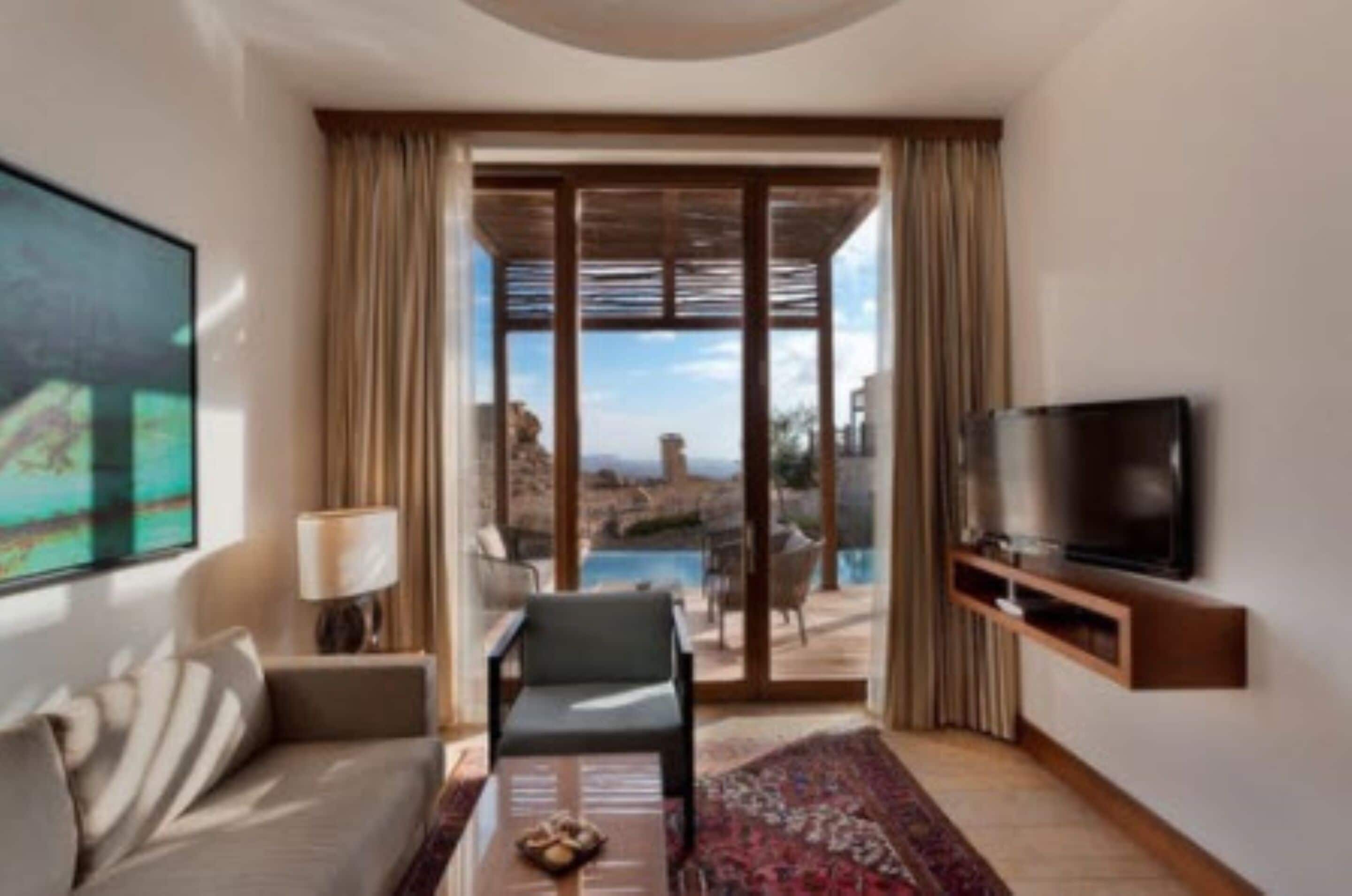 A modern hotel room with a sofa, a small table, a TV, and a view of a pool and mountains.