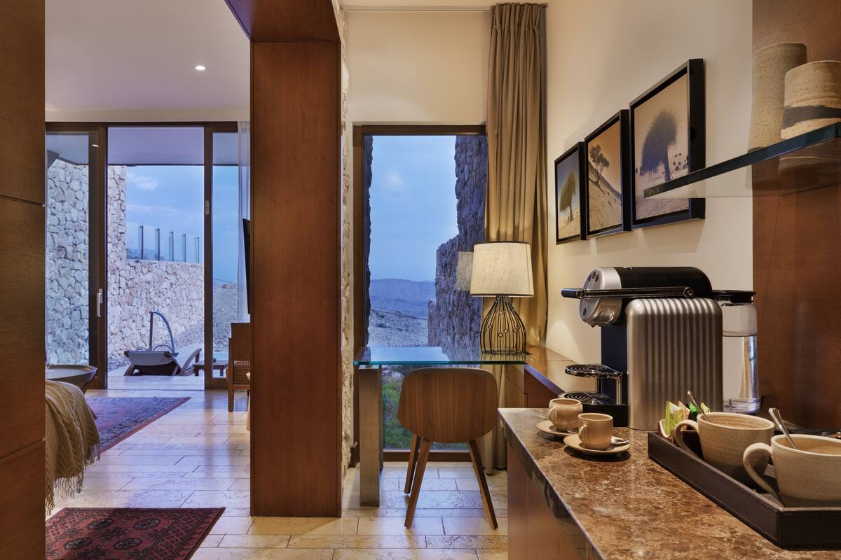 Quarto de hotel moderno com parede de pedra, porta de vidro para uma varanda, um espaço para café e vista para as montanhas.