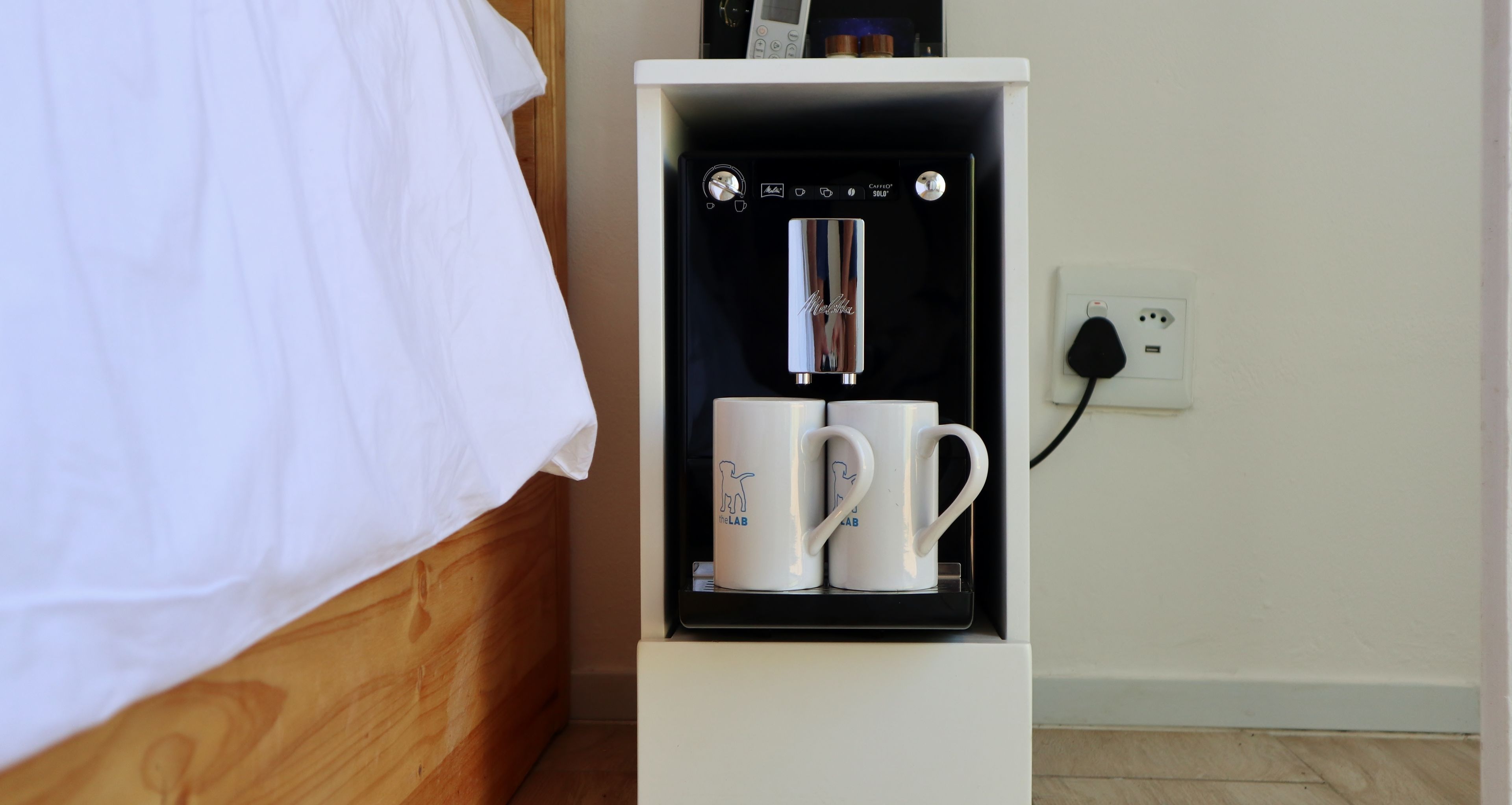 A white cabinet with a coffee machine and two white mugs labeled "The LAB" on a wooden bed.