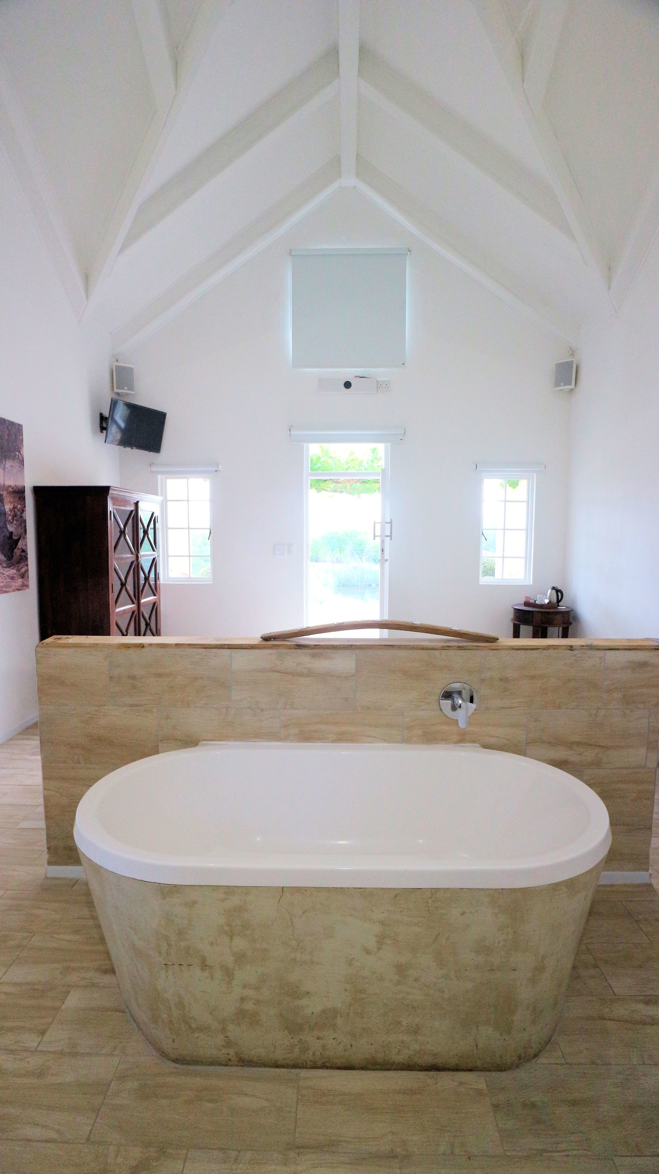 A spacious bathroom with a large freestanding bathtub, a wooden cabinet, and a television mounted on the wall.