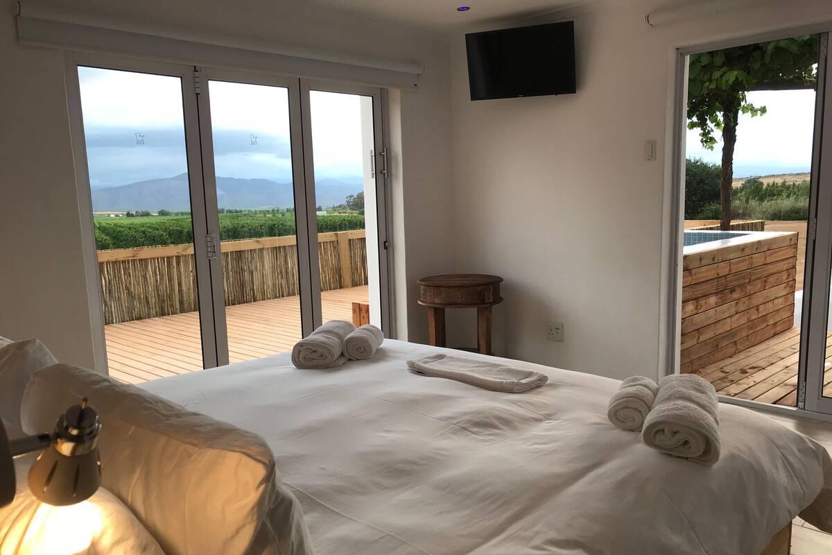 A bedroom with a bed, a bedside table, a TV, and a view of a wooden deck and greenery.