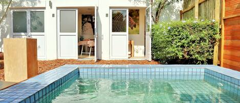 Una piscina con borde de azulejos, una terraza de madera y un edificio blanco con dos puertas abiertas.