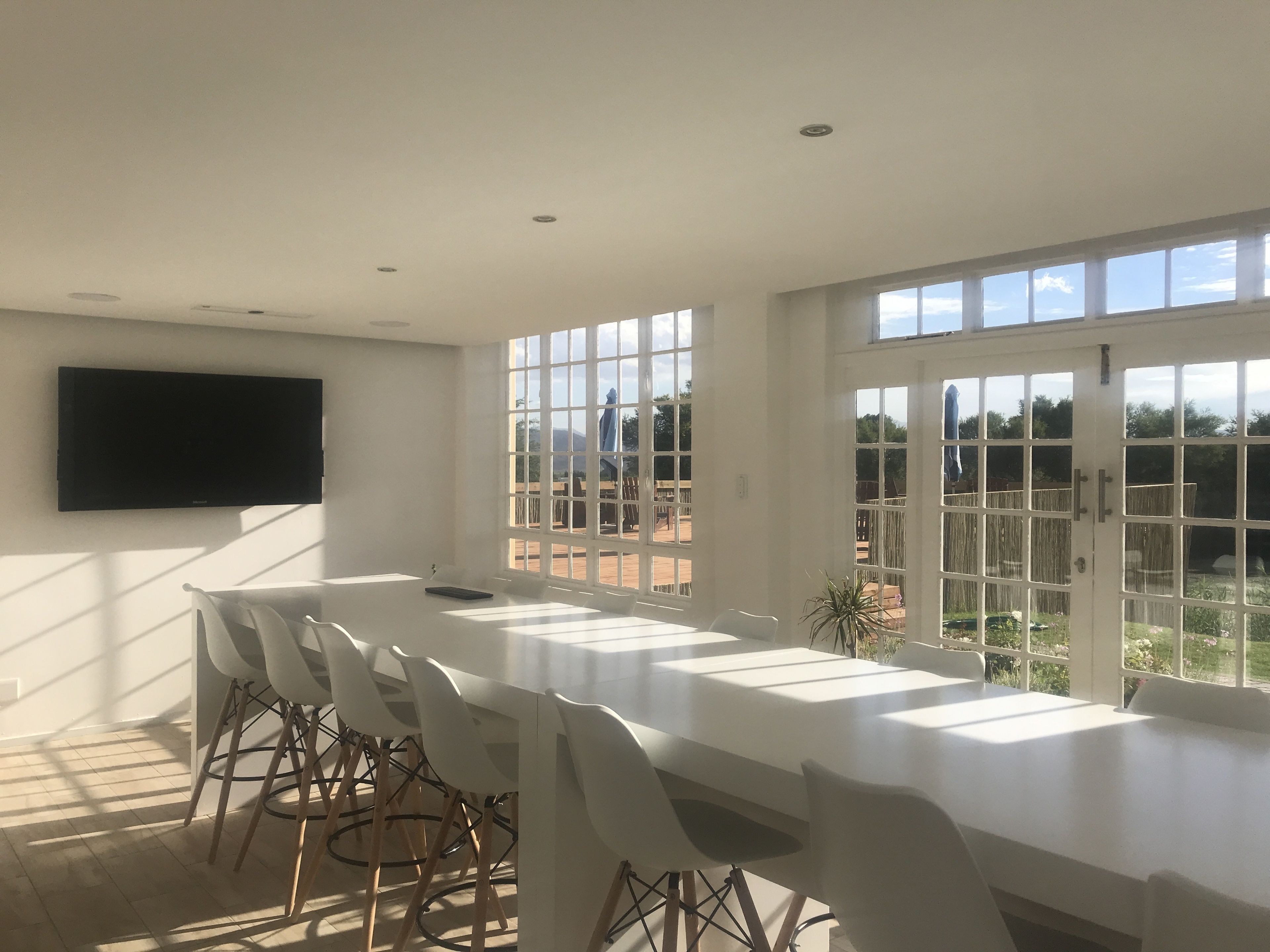 A spacious dining area with a long table and white chairs, large windows with a view of the outdoors, and a flat-screen TV mounted on the wall.