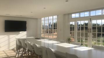 A spacious dining area with a long table and white chairs, large windows with a view of the outdoors, and a flat-screen TV mounted on the wall.