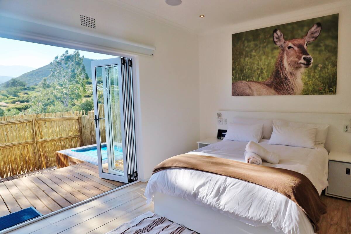 A bedroom with a bed, a nightstand, a painting of a deer, and a view of a pool and mountains.