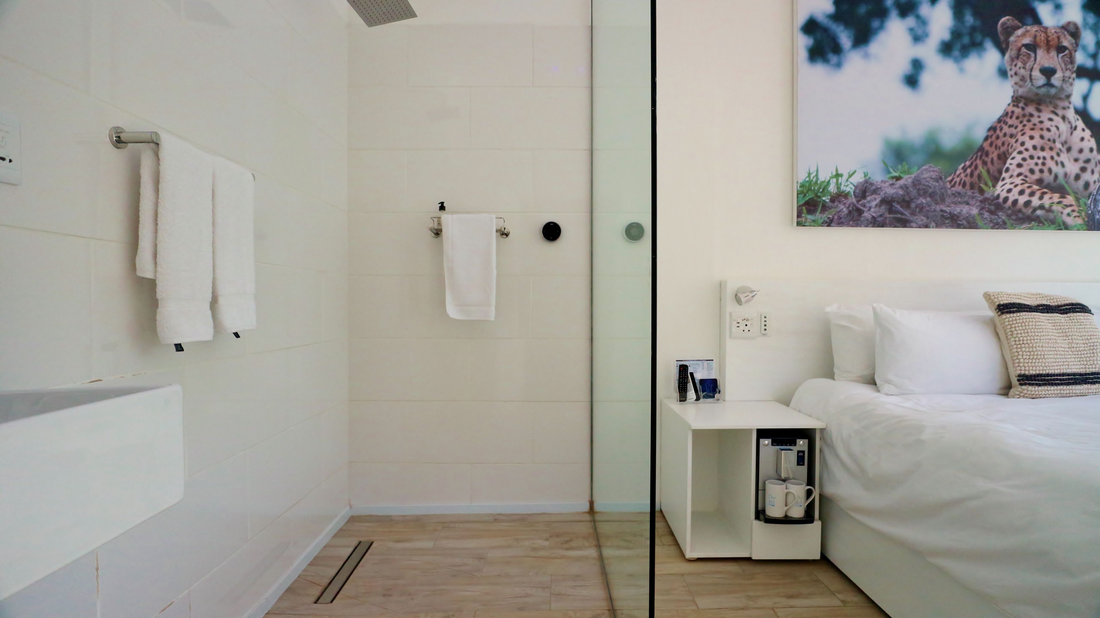 A modern bathroom with a shower, a toilet, and a sink. There is a bed with a pillow and a remote control on a nightstand.