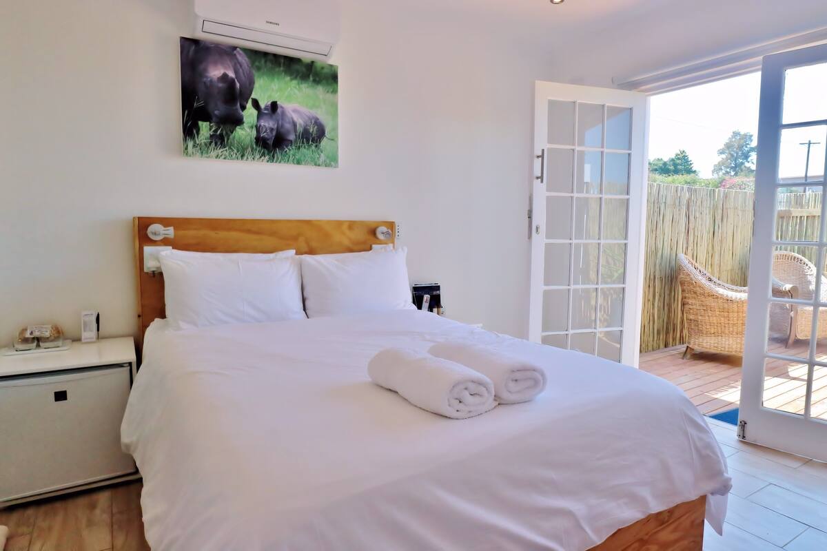 A bedroom with a large bed, white bedding, a mounted air conditioner, a nightstand, a bedside table, and a window with a view of a wooden deck and chairs.