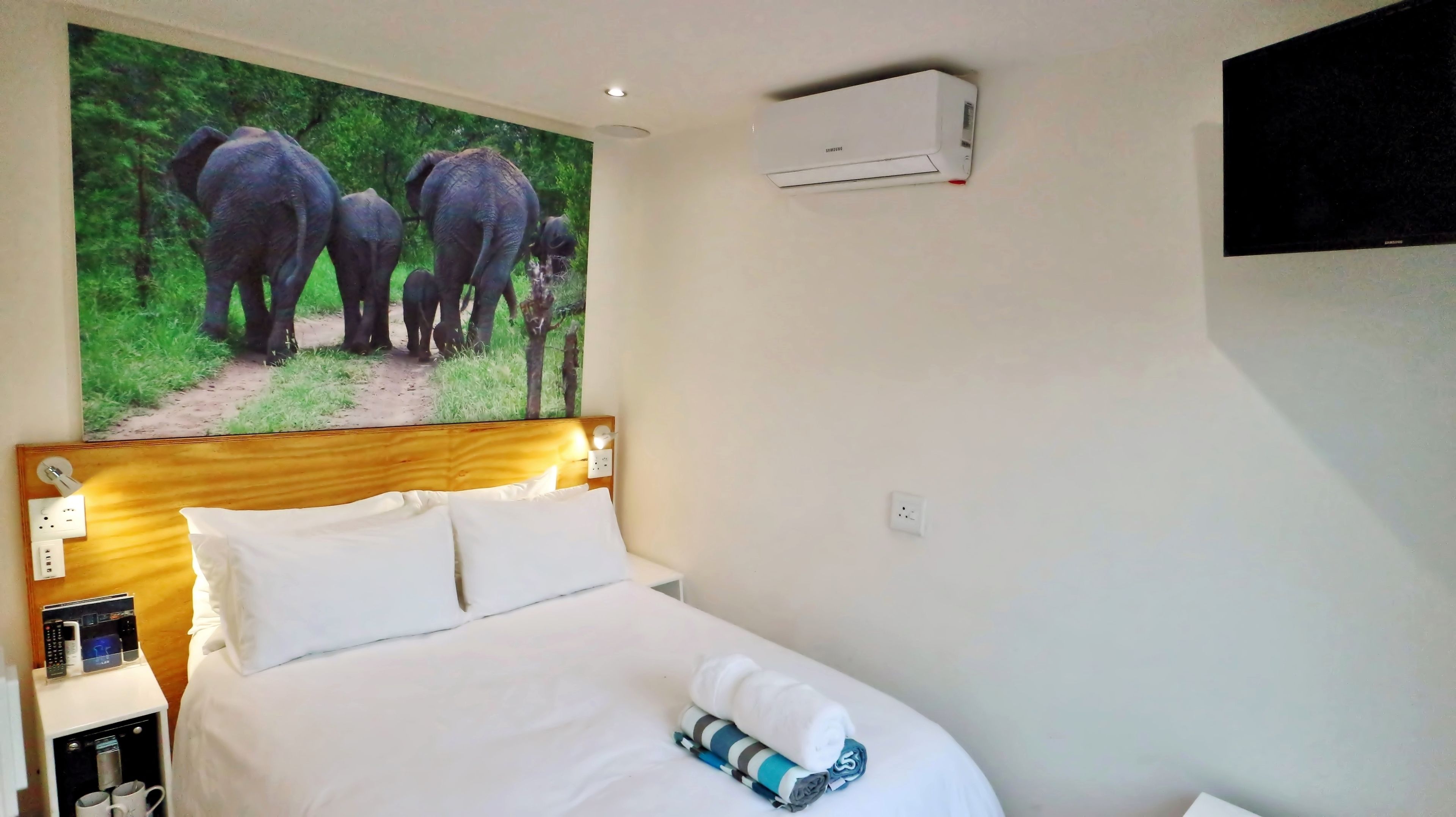 A hotel room with a bed, a wall-mounted air conditioner, a television, and a painting of elephants.