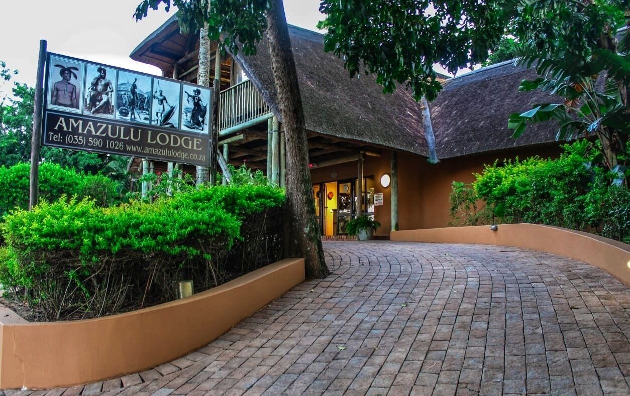 L’entrée d’Amazulu Lodge, avec un panneau indicateur, un toit de chaume et un chemin pavé menant à l’entrée.