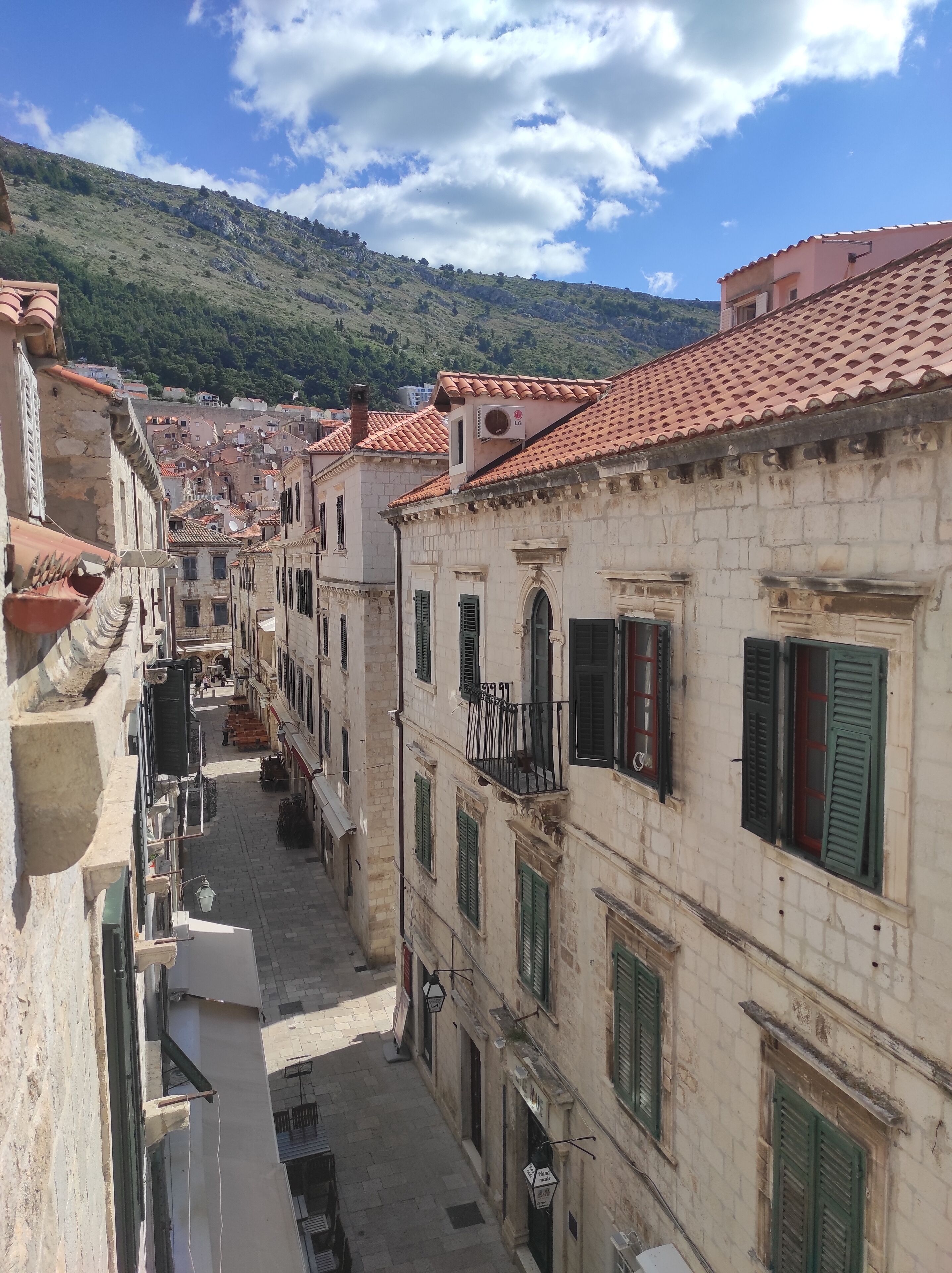 A narrow street with historic buildings, terracotta roofs, and a mountainous backdrop.