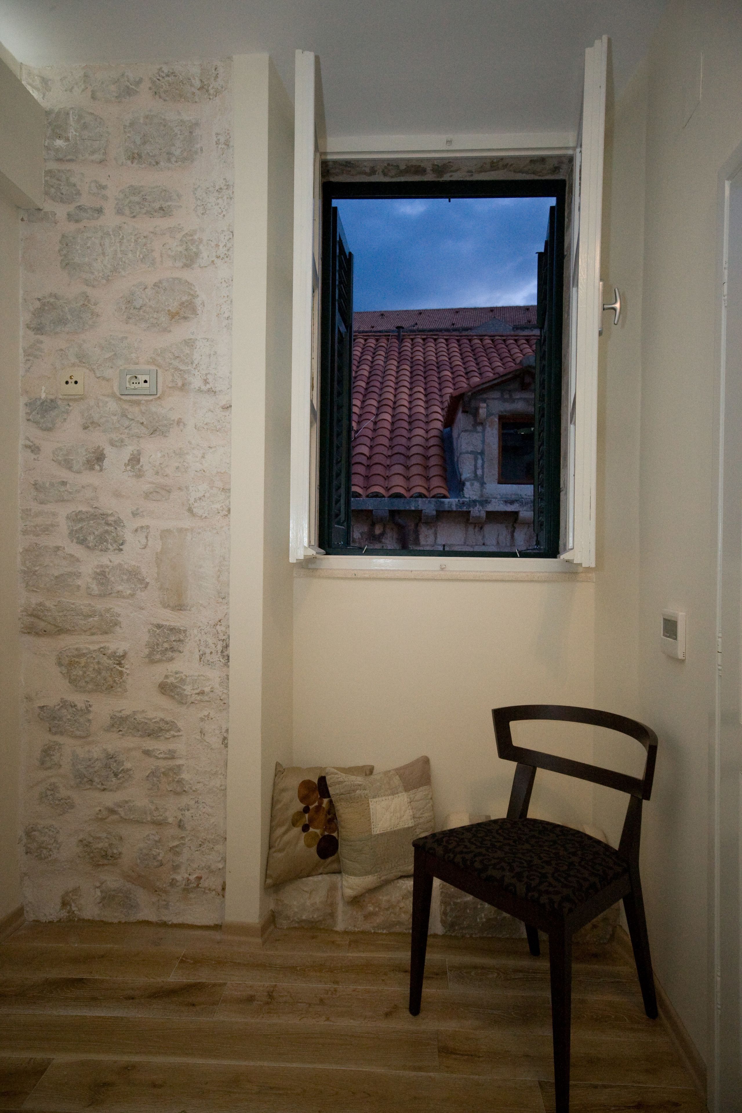Una habitación con pared de piedra, ventana con vista a un tejado de tejas rojas, una silla de madera y un cojín en el suelo.