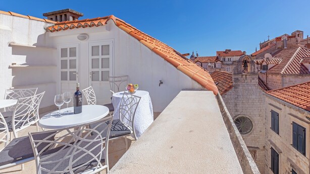 Terraço no telhado com mobiliário branco, uma mesa com garrafa e copos, e vista para a paisagem urbana.