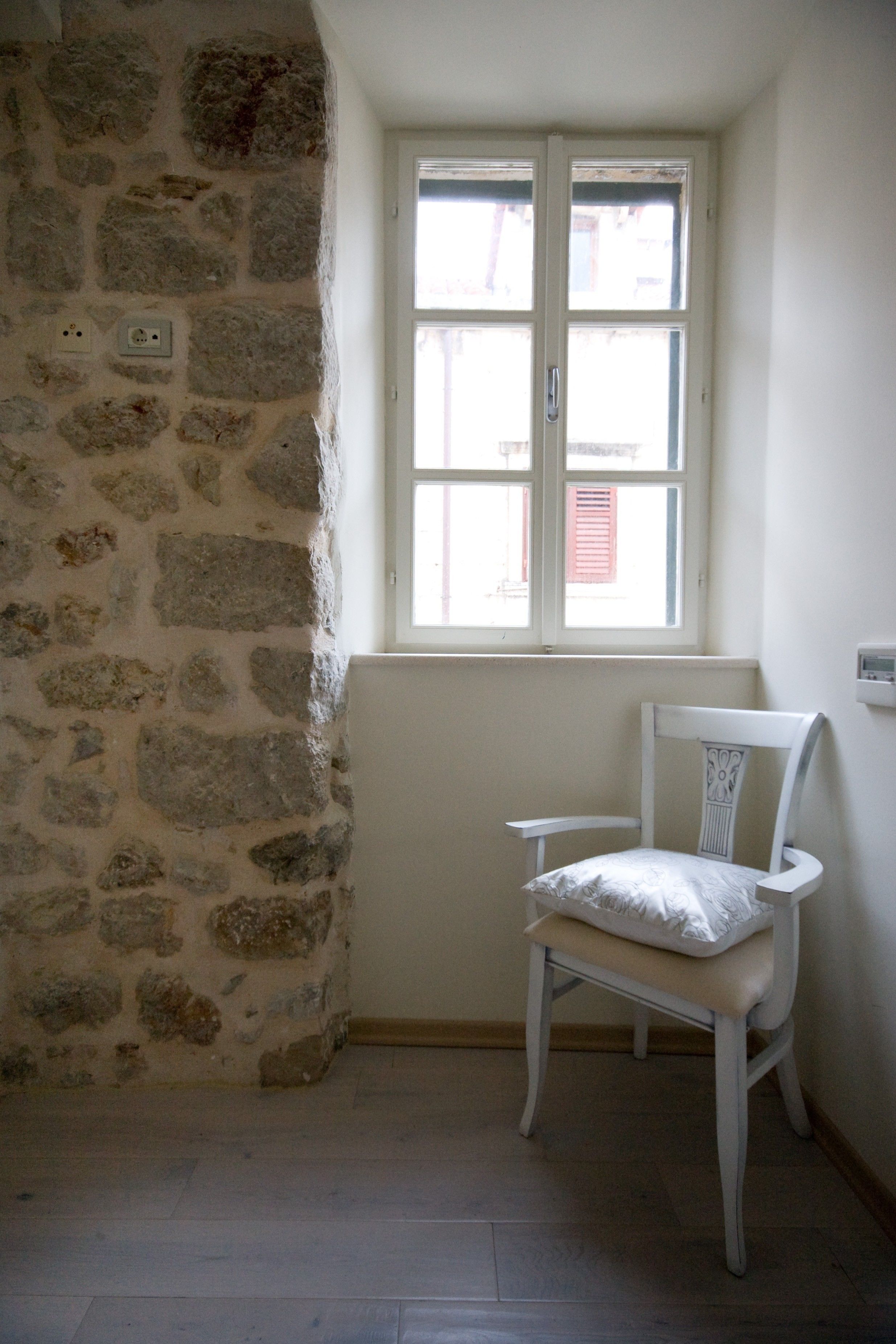 Una habitación con pared de piedra, una silla blanca, una mesita y una ventana con vistas a edificios.