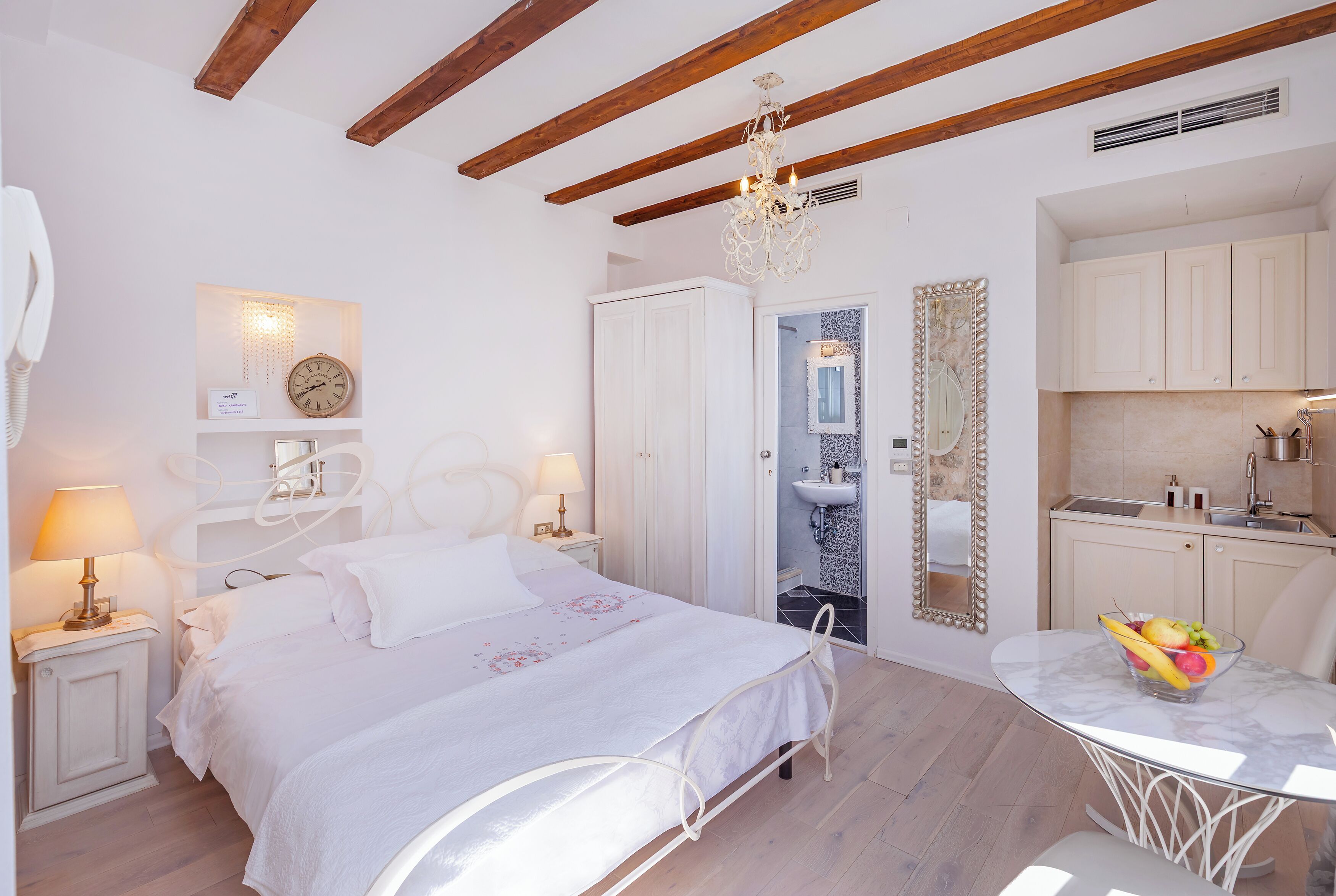A bedroom with a bed, bedside tables, a nightstand, a chandelier, a mirror, a small bathroom, and a dining area with a table and chairs.