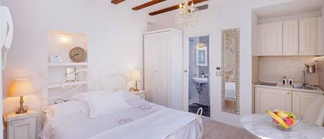 A bedroom with a bed, bedside tables, a nightstand, a chandelier, a mirror, a small bathroom, and a dining area with a table and chairs.