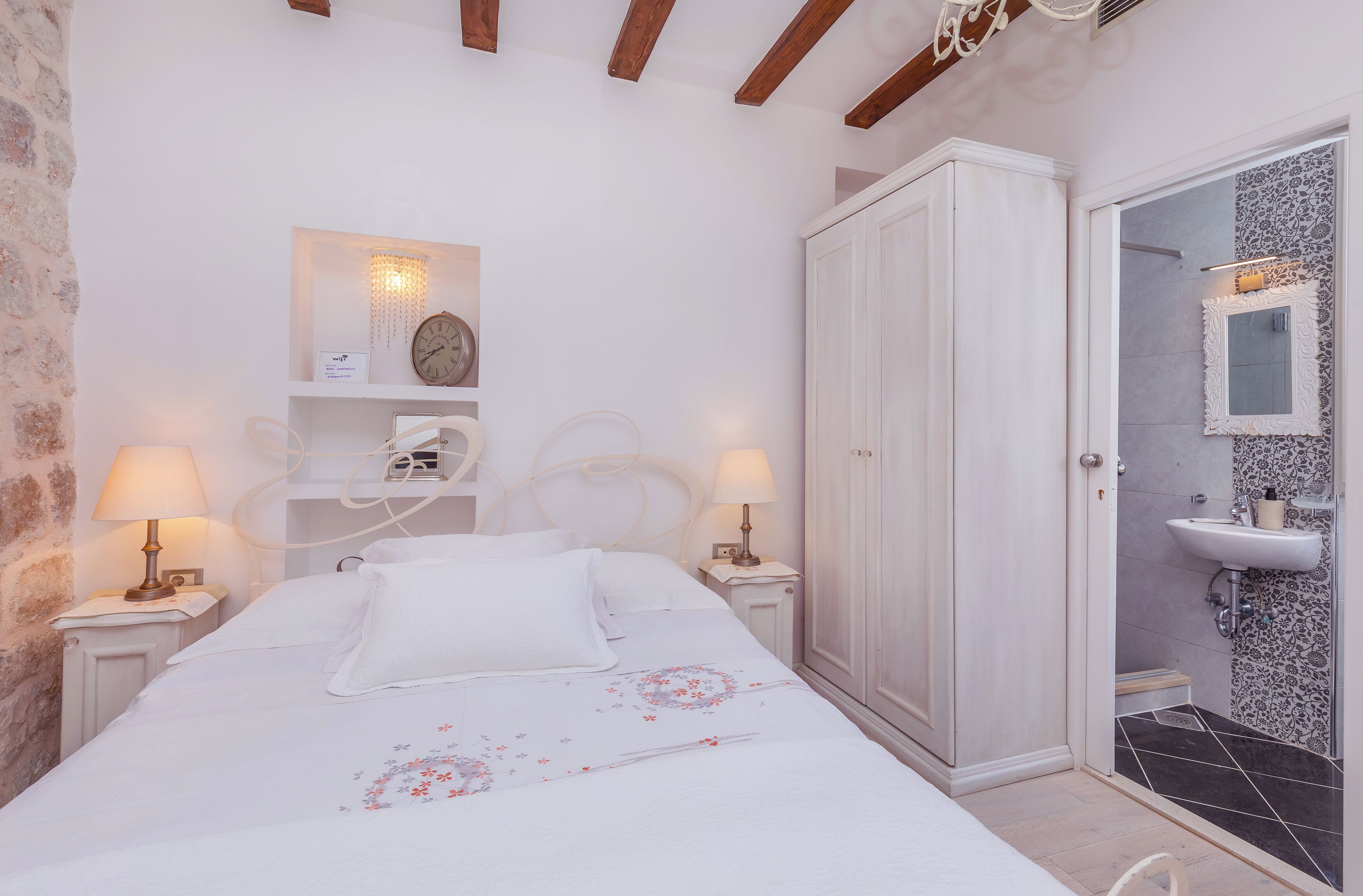 Quarto com cama, luminárias de cabeceira, guarda-roupa, mesa de cabeceira e banheiro com pia e espelho.