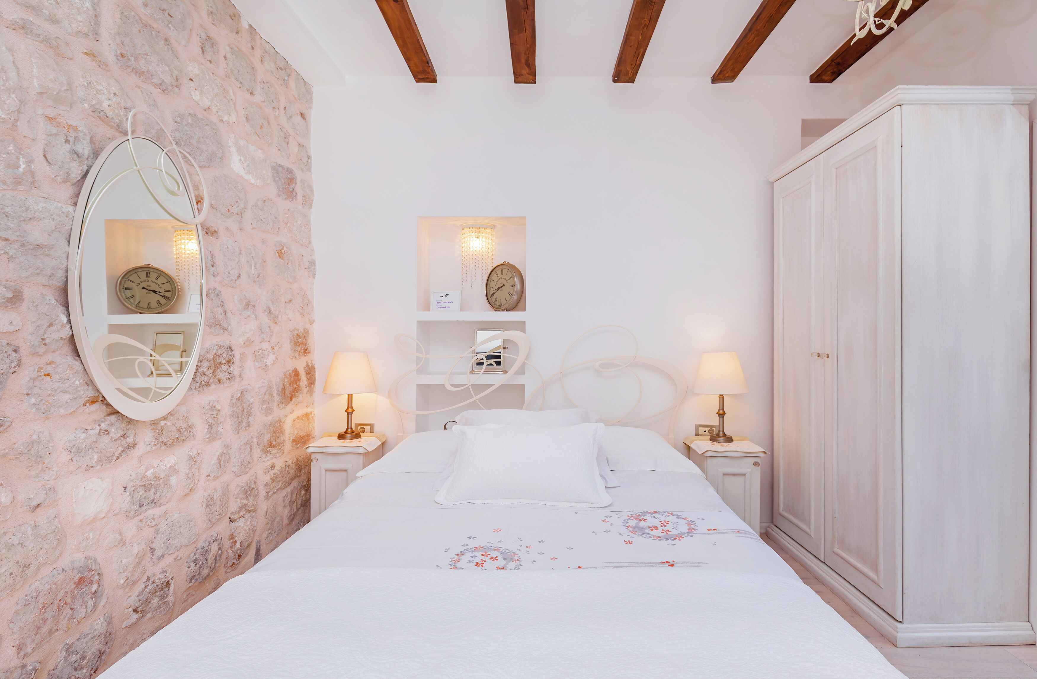 Un dormitorio con pared de piedra, una cama con ropa blanca, dos mesitas de noche con lámparas, un armario y un espejo en la pared.