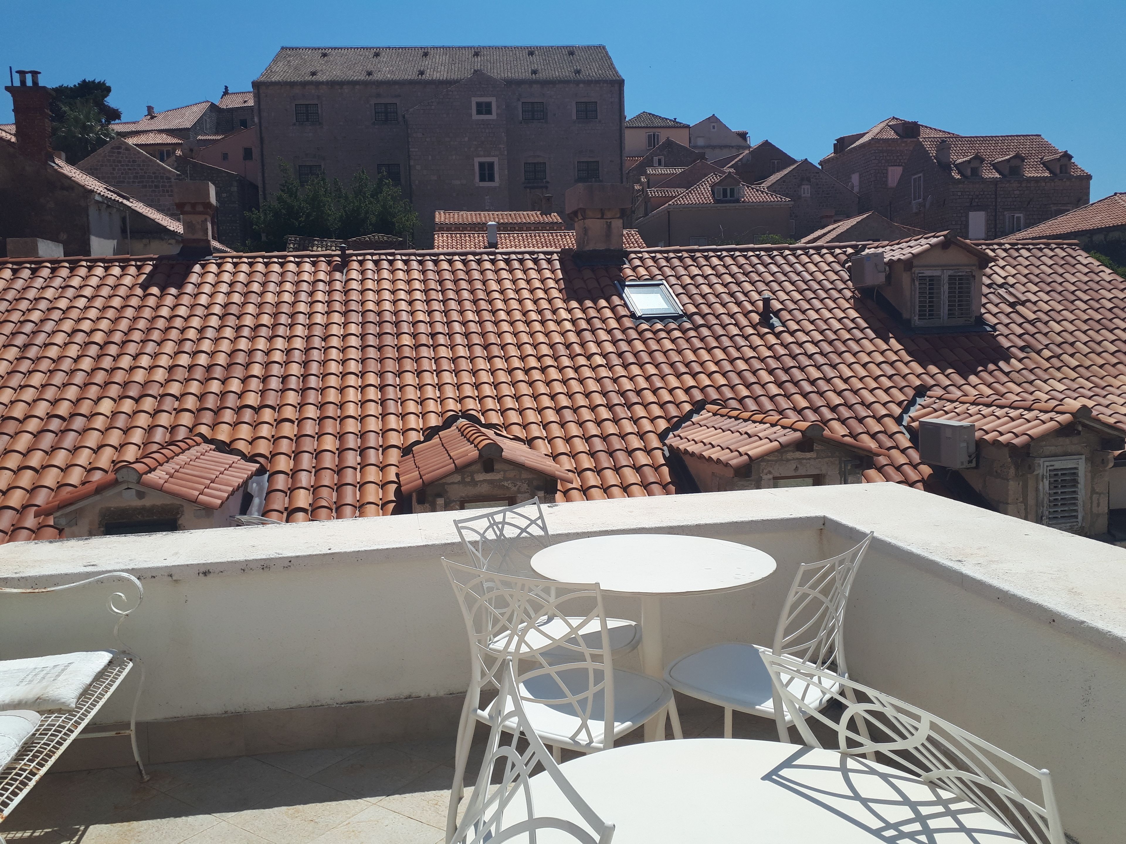 A terrace with white metal chairs and a small table, overlooking a cityscape with terracotta-tiled roofs and stone buildings.