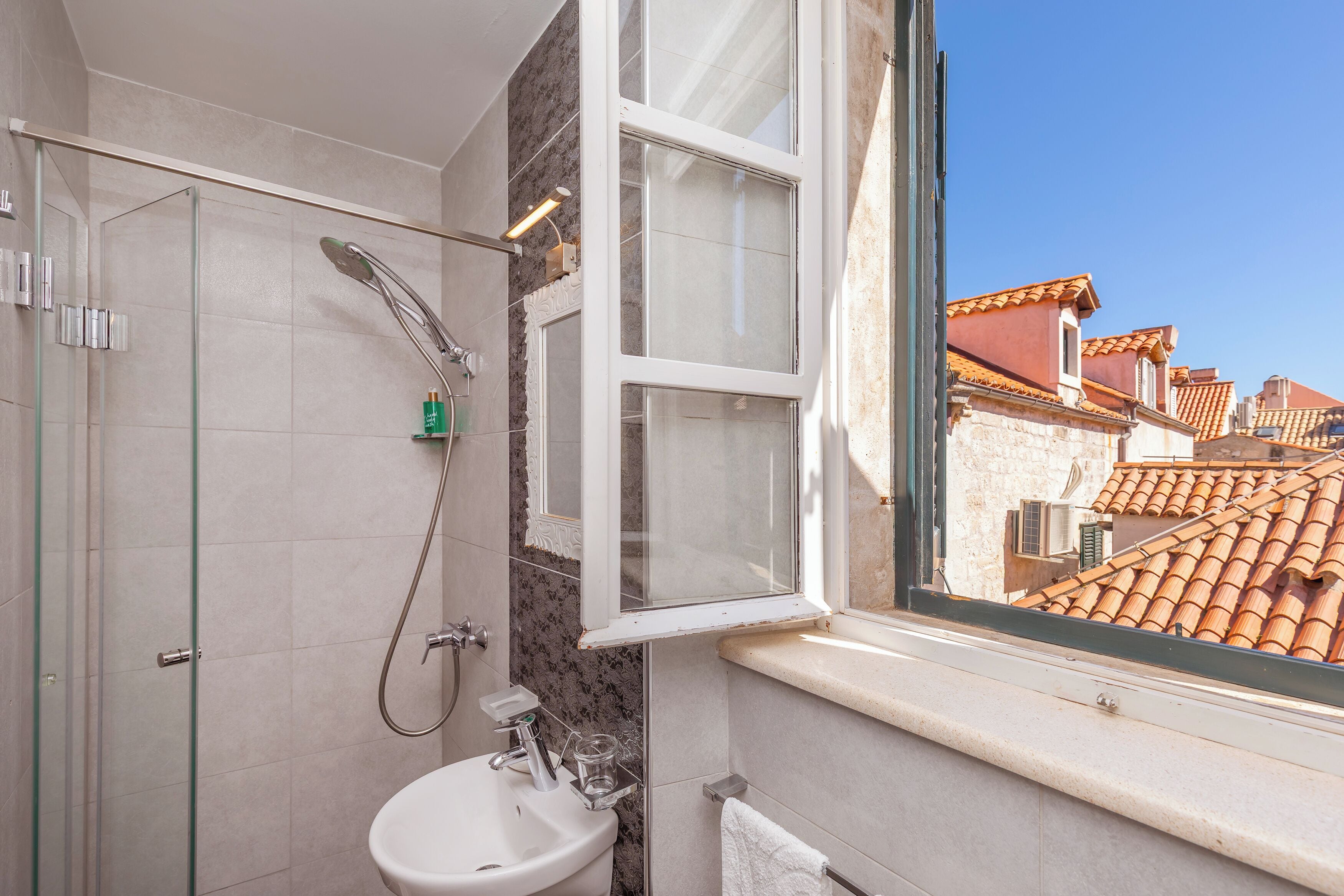 Een badkamer met een douchecabine van glas, een wastafel en een raam met uitzicht op een betegeld dak.