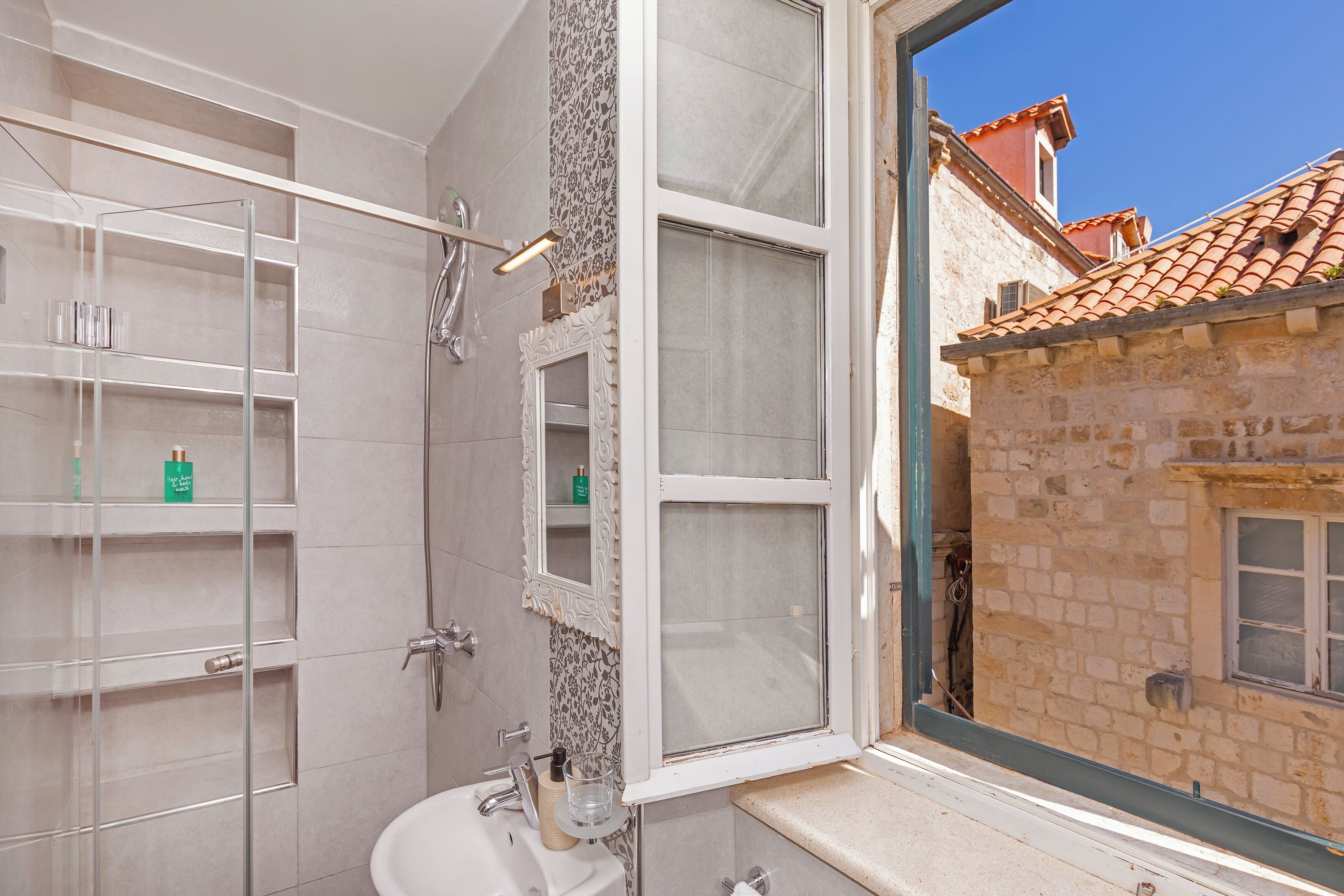 A bathroom with a glass shower enclosure, a sink, and a window overlooking a stone building.