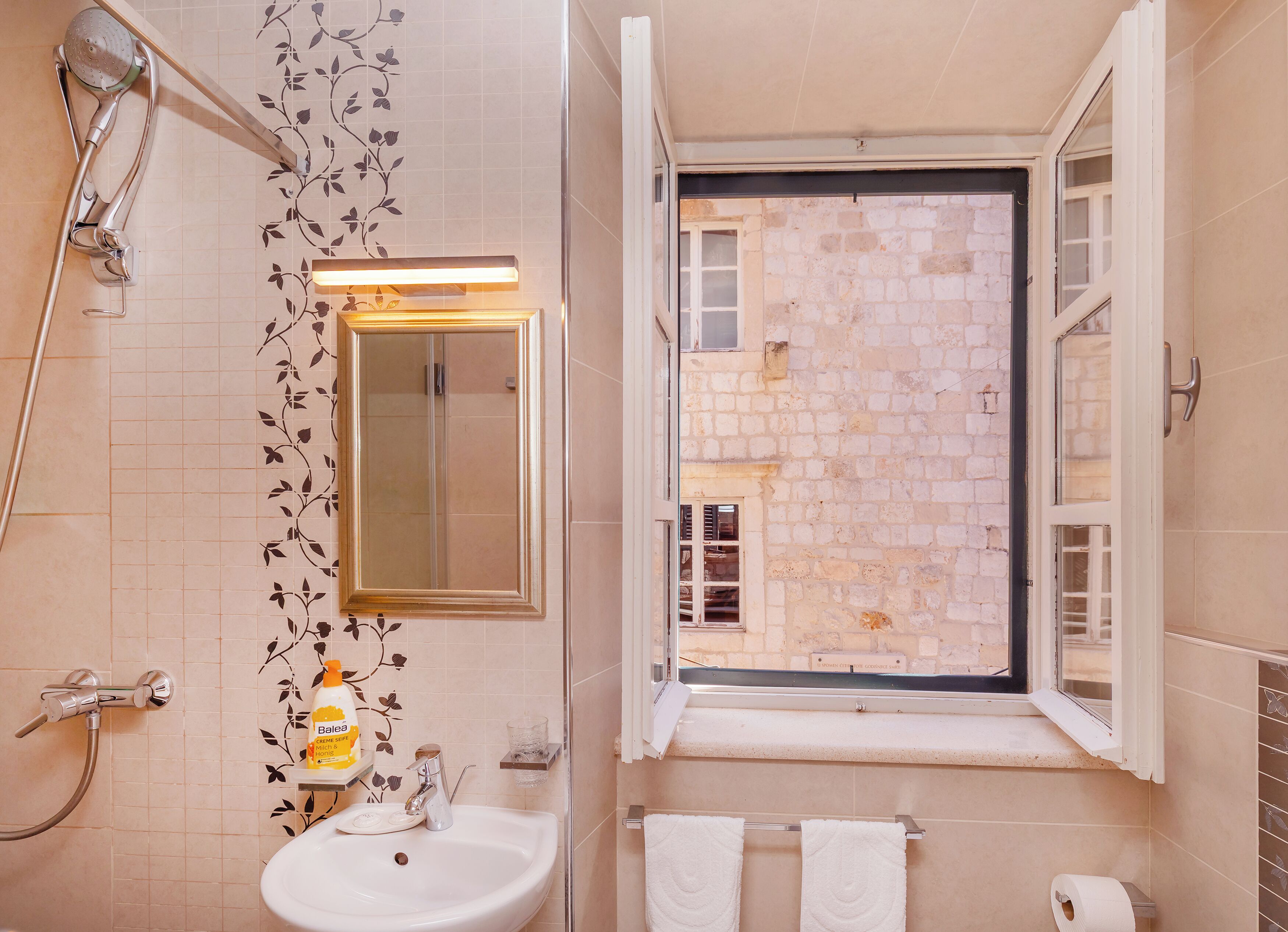 Un baño con lavabo, espejo, ducha y ventana con vistas a un edificio de piedra.