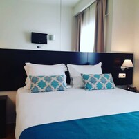 Una camera d'albergo con un letto grande, due cuscini decorativi, una televisione a schermo piatto, una lampada da comodino e una finestra con tende.