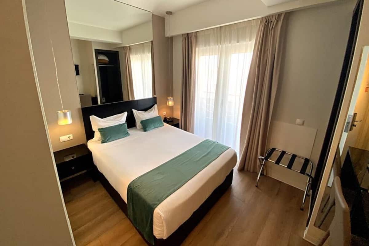 Habitación de hotel con una cama grande, una mesita de noche con lámpara, un espejo y vistas al exterior a través de cortinas.