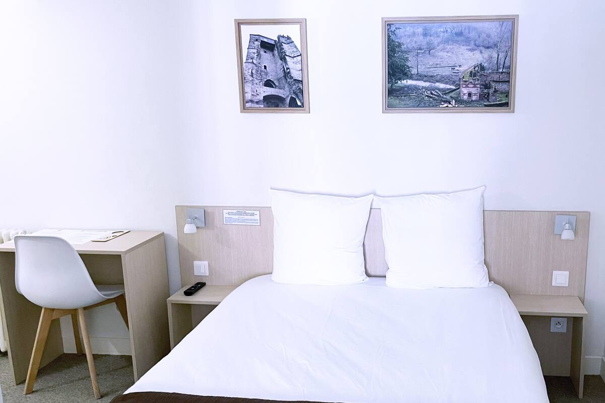 A hotel room with a bed, a desk, and two chairs. There are two framed pictures on the wall.