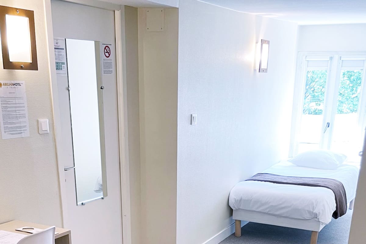 A hotel room with a desk, a chair, a bed, and a large window.