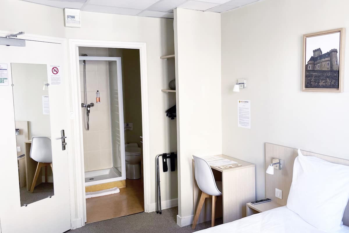A hotel room with a bed, a desk with a chair, a small table, a nightstand, a wall-mounted lamp, and a picture frame.