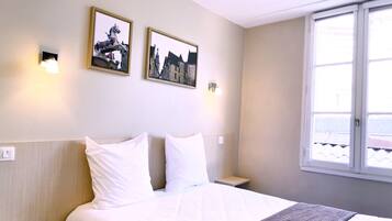 Una habitación de hotel con cama, dos almohadas, una mesita de noche, dos lámparas de pared y dos cuadros enmarcados.